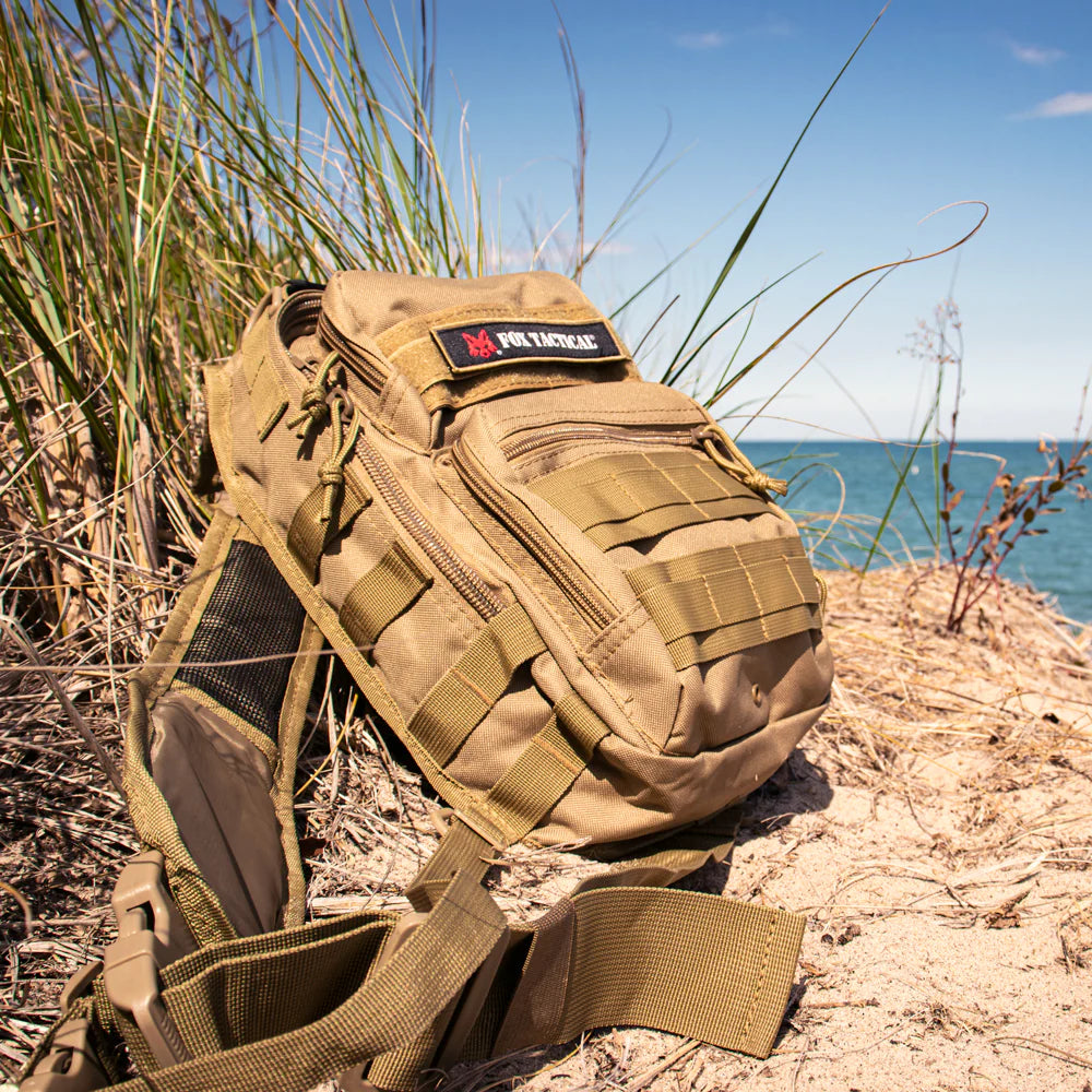Tan tactical backpack on a sandy beach with grass and water in the background