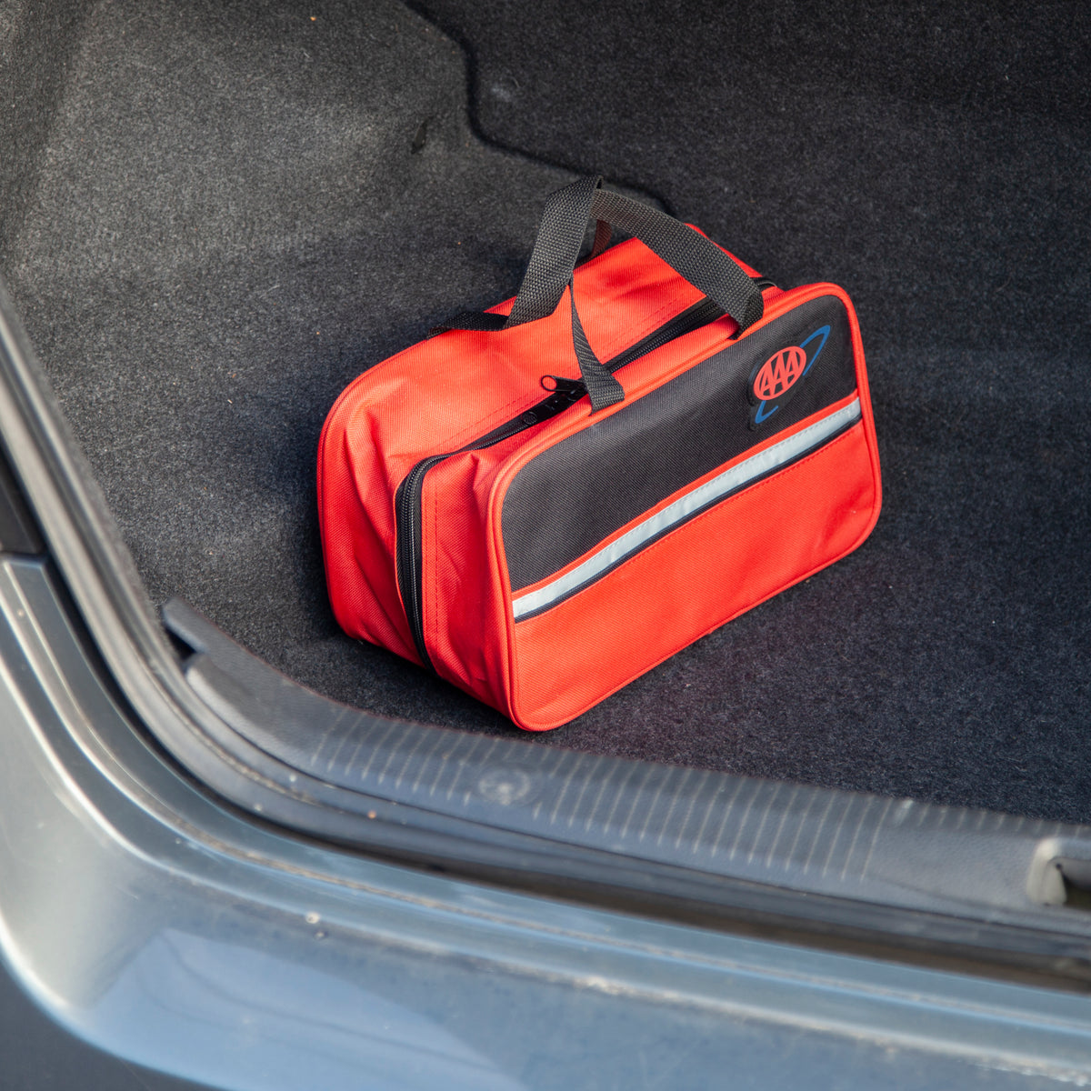 A red and black utility bag with a reflective stripe and a circular logo patch, placed inside the open trunk of a car.