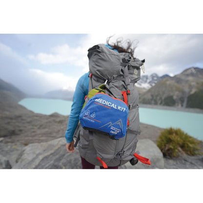 A person wearing a large gray hiking backpack stands outdoors near a turquoise lake and mountains, with a blue and red Medical Kit pouch attached to the back of the pack.