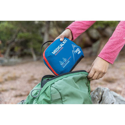 Person holding a blue First Aid Kit next to a green bag outdoors.