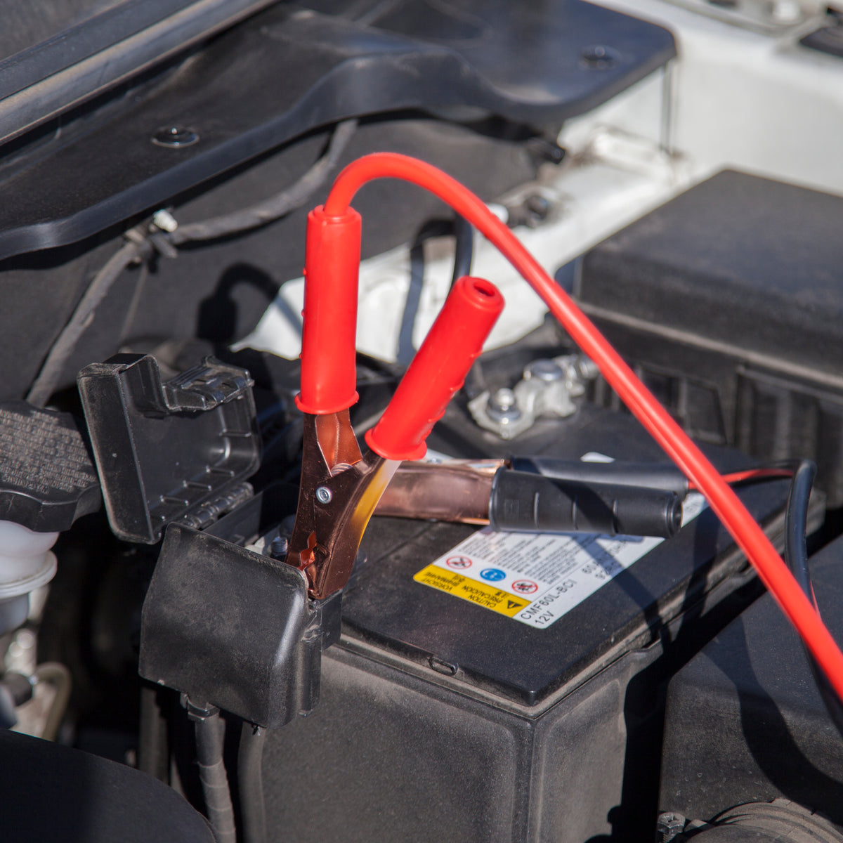 A car battery with red jumper cables attached in a close-up view of an open engine bay.
